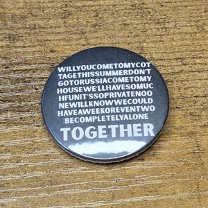 May include: A round, black button with white text. The text includes the words "TOGETHER" and a series of phrases. The button is on a wooden surface.