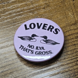 May include: A round, light purple button with the word "LOVERS" in black, bold letters at the top. Below are two black loon bird illustrations facing each other. The text "NO. ILYA. THAT'S GROSS." is printed in black around the bottom.
