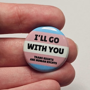 Op de afbeelding: Een ronde button met een roze, witte en lichtblauwe kleur. De button heeft de tekst "I'LL GO WITH YOU" en "TRANS RIGHTS ARE HUMAN RIGHTS" in zwarte letters. De button wordt tussen twee vingers gehouden.