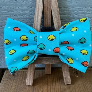 May include: A turquoise bow tie with a repeating pattern of cartoon fish in yellow, orange, and green. White polka dots are scattered across the fabric. The bow tie is displayed on a small wooden easel.