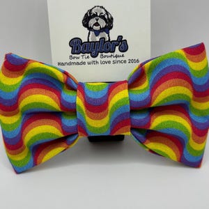 May include: A colorful rainbow-striped bow tie. The bow tie has a wavy pattern of red, orange, yellow, green, blue, and purple. A white tag with the logo "Baylor's Bow Tie Boutique" is attached.