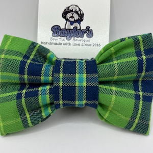 May include: A green and navy blue plaid bow tie. The bow tie is attached to a white tag with the text "Baylor's Bow Tie Boutique Handmade with love since 2016" and a logo of a dog wearing a bow tie.