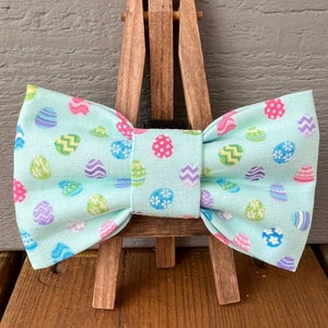 May include: A light blue bow tie with a repeating pattern of colorful Easter eggs. The eggs are decorated with various designs and colors, including pink, purple, blue, and green. The bow tie is made of fabric and is displayed on a small wooden easel.