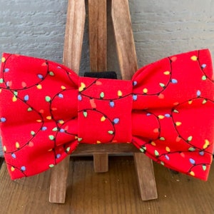 May include: Red bow tie with a pattern of colorful Christmas lights. The bow tie is displayed on a wooden easel.