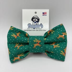 May include: A green bow tie featuring a repeating pattern of tan reindeer with red collars and white polka dots. The bow tie is attached to a white display card with a logo of a dog and the text Bayder's Bow Tie Boutique Handmade with love since 2016.
