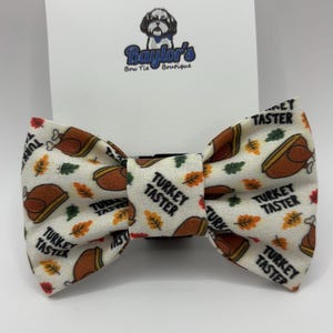 May include: A cream-colored bow tie with a repeating pattern of roasted turkeys and autumn leaves. The bow tie has the text "TURKEY TASTER" printed on it. The bow tie is from Baylor's Bow Tie Boutique.