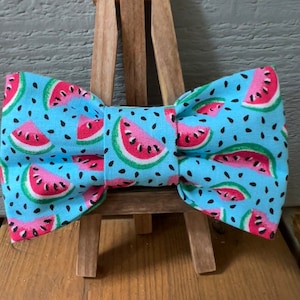 May include: A blue bow tie with a watermelon print. The bow tie is tied and displayed on a wooden easel.
