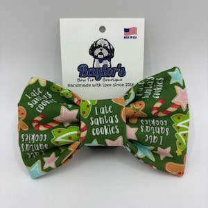 May include: A green fabric bow tie featuring a repeating pattern of gingerbread men, candy canes, and stars, with the text I ate Santa's cookies printed repeatedly. The bow tie is attached to a white display card with a dog logo and text.
