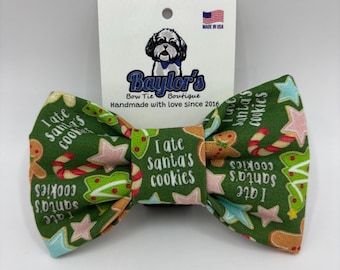 May include: A green fabric bow tie featuring a repeating pattern of gingerbread men, candy canes, and stars, with the text I ate Santa's cookies printed repeatedly. The bow tie is attached to a white display card with a dog logo and text.
