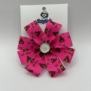 May include: A bright pink dog bow with a flower design. The bow features a repeating pattern of a brown poop emoji and the words "lil shit". The center of the bow has a silver rhinestone embellishment.