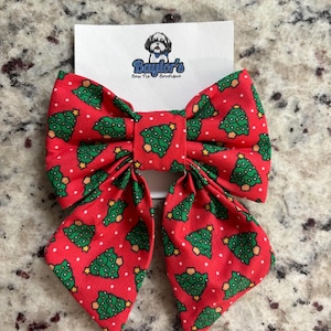 May include: Red bow tie with a Christmas tree pattern. The bow tie is made of fabric and has a white polka dot pattern. The bow tie is tied in a bow shape and has a small tag that says "Baylor's Bow Tie Boutique".