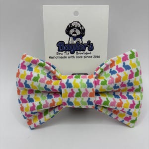 May include: A colorful bow tie with a repeating pattern of bunny silhouettes in pink, blue, yellow, and green on a white background. The bow tie is centered in front of a white tag with the text "Baylor's Bow Tie Boutique Handmade with love since 2016".