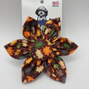 May include: A fabric flower accessory featuring a brown background with a pattern of orange, yellow, and green autumn leaves and small white dots. The flower has six pointed petals and a circular center, attached to a white display card with a dog logo.