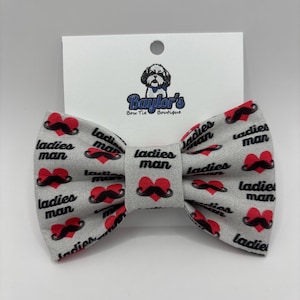 May include: A gray bow tie with a repeating pattern of red hearts with black mustaches and the words "ladies man". The bow tie has a white tag with the logo "Baylor's Bow Tie Boutique".