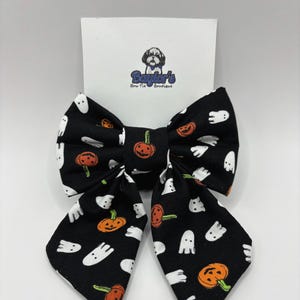May include: A black Halloween-themed bow tie featuring a repeating pattern of white ghosts and orange jack-o'-lanterns. The bow tie is centered on a white card with the logo "Baylor's Bow Tie Boutique" and a dog illustration.