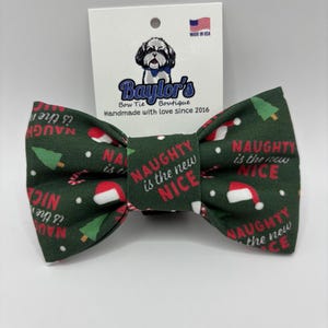 May include: A dark green bow tie featuring a repeating Christmas pattern of Santa hats, candy canes, and pine trees, with the text "NAUGHTY is the new NICE" printed in red and white. The bow tie is attached to a white branded display card.