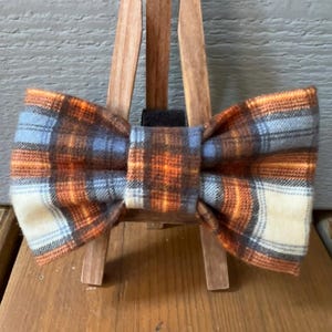 May include: A plaid bow tie in orange, blue, and white. The bow tie is tied and displayed on a wooden easel.