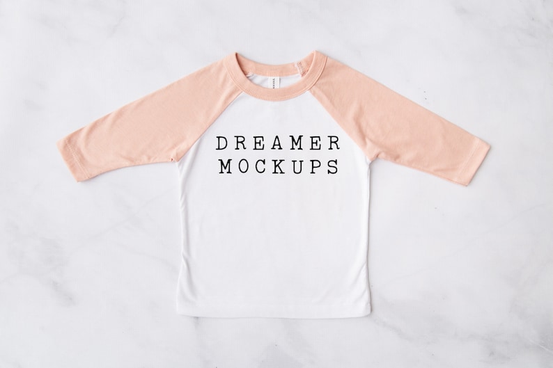 May include: A white long-sleeved t-shirt with peach colored sleeves. The shirt has a baseball style design and the text "DREAMER MOCKUPS" printed on the front.
