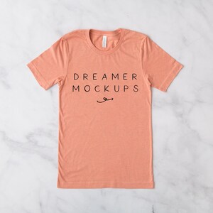 May include: A coral coloured t-shirt with short sleeves and a crew neck. The shirt has the text "DREAMER MOCKUPS" printed on the front in black.