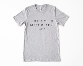 May include: A light gray t-shirt with the text "DREAMER MOCKUPS" in black, centered on the front. A small curved arrow is below the text. The shirt has short sleeves and a crew neck.