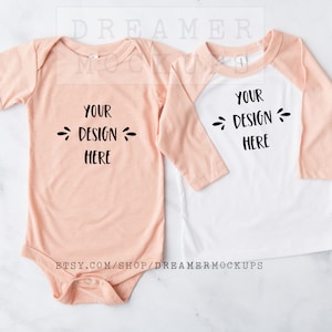 May include: Two baby shirts, one a peach-colored short-sleeved bodysuit and the other a white long-sleeved baseball tee with peach-colored sleeves. Both shirts have the text "YOUR DESIGN HERE" printed on them.