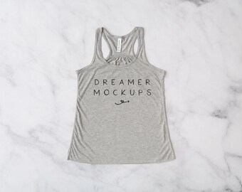 May include: A heather gray racerback tank top with the text "DREAMER MOCKUPS" in black, and a small curved arrow design. The top is made of a soft material and is displayed on a white marble surface.