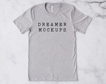 May include: A heather gray t-shirt with the words "DREAMER MOCKUPS" printed in black, centered on the front. The shirt is a crew neck style and is displayed on a white marble surface.