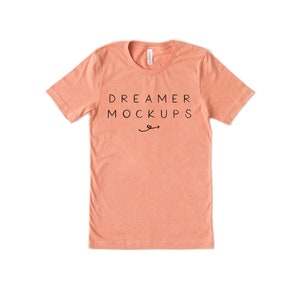 May include: A peach-coloured t-shirt with short sleeves and a crew neck. The shirt has the text "DREAMER MOCKUPS" printed on the front in black.