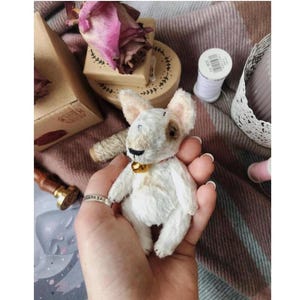 May include: A white and brown teddy bear shaped like a bull terrier, wearing a red collar with a gold bell. The teddy bear is 10 cm tall and is being held in someone's hand. The background is a pink and white checked fabric.