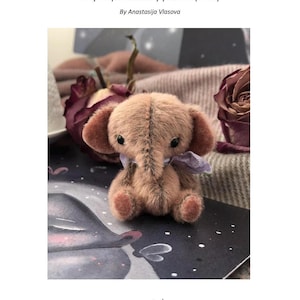 以下が含まれることがあります： 首に紫のリボンを巻いた、薄い茶色の小さなぬいぐるみゾウ。ぬいぐるみは、白い星が散りばめられた暗い背景の上に座っています。ぬいぐるみの上には、「Tiny elephant teddy pattern (5 cm)」と「By Anastasija Vlasova」の文字があります。