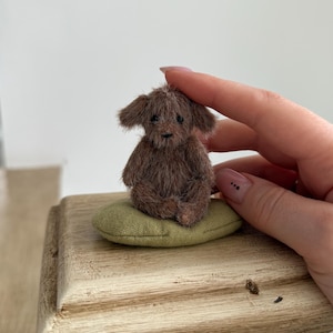 May include: A miniature brown plush dog figurine with floppy ears sits on a small, olive green pillow. The tiny dog is perched on a wooden surface. The figurine is a handcrafted item.