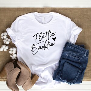 May include: White t-shirt with the text "Flattie Baddie" in black script with a small black heart.