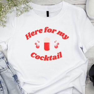 Puede incluir: Camiseta blanca con el texto rojo "Here for my Cocktail" y un gráfico de cóctel. La camiseta está sobre una superficie clara con vetas de madera, con vaqueros azules, zapatillas blancas y auriculares negros.