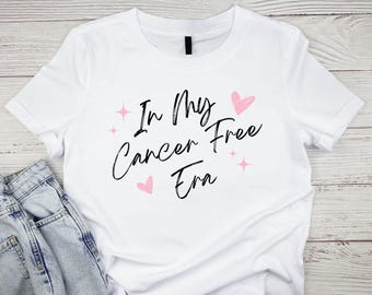 Peut inclure: T-shirt blanc avec le texte "In My Cancer Free Era" en écriture noire avec des cœurs roses et des accents de paillettes.