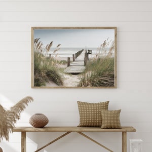 Könnte beinhalten: Ein gerahmtes Foto eines Holzstegs, der zu einem Steg an einem Sandstrand führt. Der Strand ist von hohem Gras umgeben und das Meer befindet sich im Hintergrund.