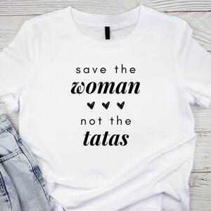 May include: White t-shirt with black text that reads "save the woman not the tatas" with three small hearts above the word "woman".