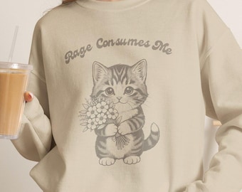 Op de afbeelding: Een beige sweater met ronde hals en een donkergrijze afbeelding van een cyperse kitten die een boeket bloemen vasthoudt. Boven de kitten staat de tekst "Rage Consumes Me" gedrukt in een gebogen, retro-stijl lettertype.