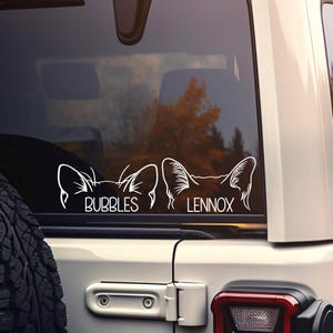 May include: A white vinyl decal applied to a vehicle rear window, featuring two sets of stylized animal ears in outline form. Below the left set of ears is the name BUBBLES, and below the right set of ears is the name LENNOX in white lettering.