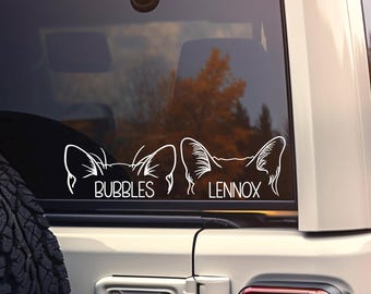 May include: A white vinyl decal applied to a vehicle rear window featuring two sets of stylized animal ears. Below the left set of ears is the name BUBBLES in white capital letters, and below the right set of ears is the name LENNOX in white.
