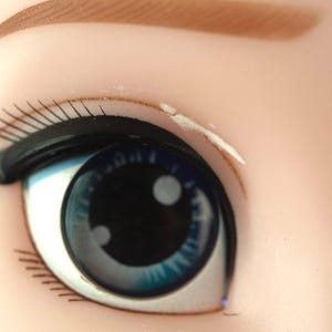 K&ouml;nnte beinhalten: Nahaufnahme eines Puppenauges. Das Auge ist leuchtend blau mit einer schwarzen Pupille und einem wei&szlig;en Glanzlicht. Schwarzer Eyeliner und lange, schwarze Wimpern umrahmen das Auge. Die Augenbraue ist hellbraun.