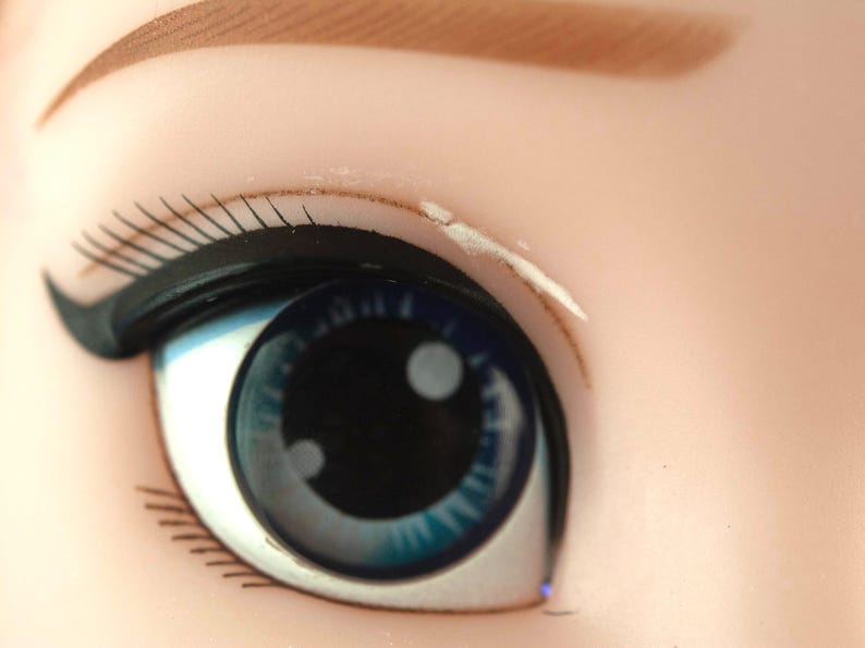 K&ouml;nnte beinhalten: Nahaufnahme eines Puppenauges. Das Auge ist leuchtend blau mit einer schwarzen Pupille und einem wei&szlig;en Glanzlicht. Schwarzer Eyeliner und lange, schwarze Wimpern umrahmen das Auge. Die Augenbraue ist hellbraun.