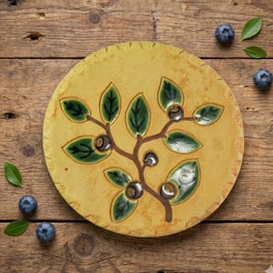 May include: A round, yellow ceramic plate with a hand-painted design of a blueberry branch with green leaves and dark blue berries. The plate is surrounded by fresh blueberries and leaves on a wooden surface.