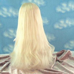 May include: A long, light blonde wig with wavy ends. The wig is styled to fall down the back, with a slight curl at the ends. The background is a blue fabric with white cloud patterns, and the wig rests on a pink fabric.