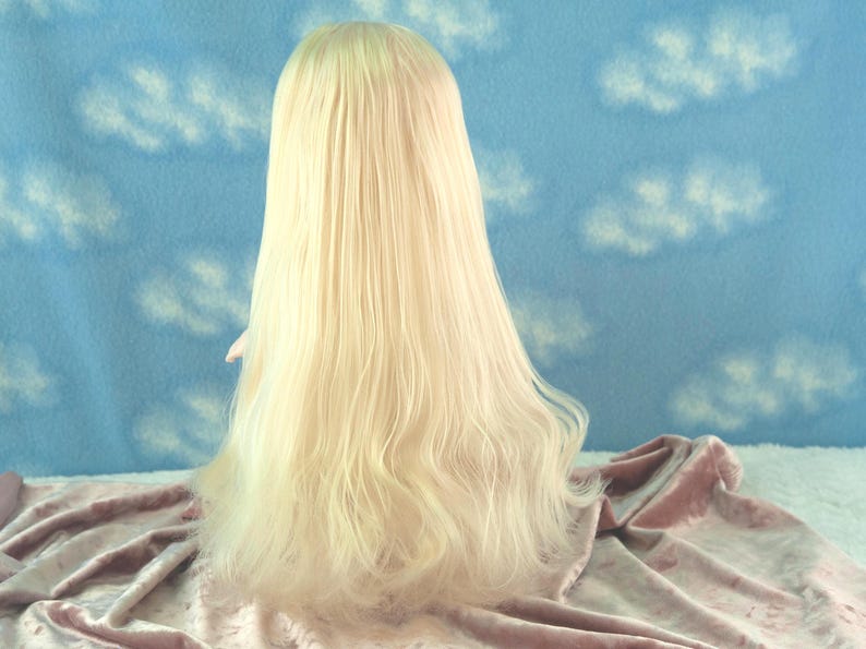 May include: A long, light blonde wig with wavy ends. The wig is styled to fall down the back, with a slight curl at the ends. The background is a blue fabric with white cloud patterns, and the wig rests on a pink fabric.