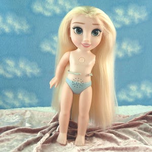 K&ouml;nnte beinhalten: Eine Puppe mit langen blonden Haaren und blauen Augen, die einen hellblauen Unterteil tr&auml;gt. Die Puppe steht vor einem blauen Hintergrund mit wei&szlig;en Wolkenmustern. Die Haut der Puppe ist hellpfirsichfarben.