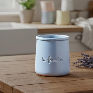 May include: A light blue ceramic jar with the embossed text "la fermière" on the front. The jar has a rounded base and a slightly flared top. It sits on a wooden surface, with other jars in the background.