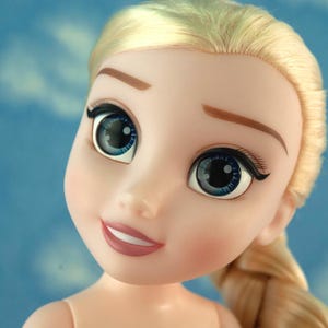 May include: Close-up of a doll's face with blonde hair styled in a side braid. The doll has large blue eyes with black eyelashes and eyebrows. The doll's skin is pale, and it has a slight smile.