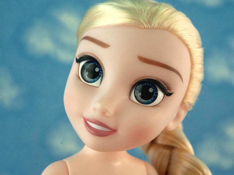 May include: Close-up of a doll's face with blonde hair styled in a side braid. The doll has large blue eyes with black eyelashes and eyebrows. The doll's skin is pale, and it has a slight smile.