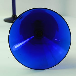 Pode incluir: Uma vista de perto de um copo de vinho azul cobalto. O copo tem uma taça larga e uma haste preta. O copo é feito de um material brilhante e reflexivo. A imagem é tirada de um ângulo baixo, olhando para dentro da taça do copo.