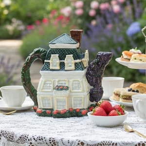 Peut inclure: Une théière en céramique fantaisiste en forme de maison, avec un toit vert, des murs blancs et une cheminée marron. La poignée est conçue pour ressembler à une branche d'arbre. La théière est posée sur une nappe en dentelle, avec un bol de fraises et des tasses à thé.
